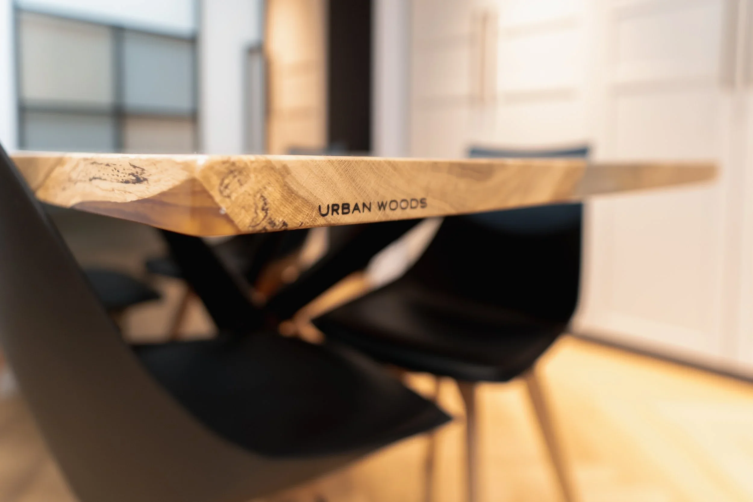 Close-up of a wooden table with the words 'URBAN WOODS' etched into the side, with black chairs around it in a modern or contemporary setting.