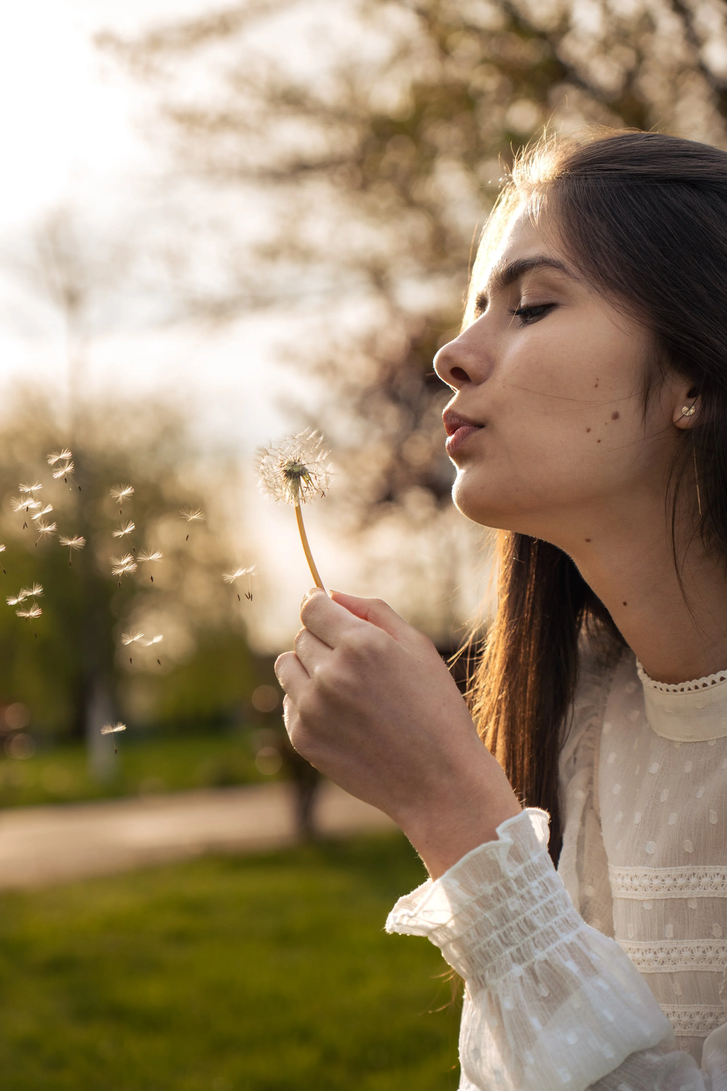 A young woman blowing on a dandelion puff in a park during sunset.