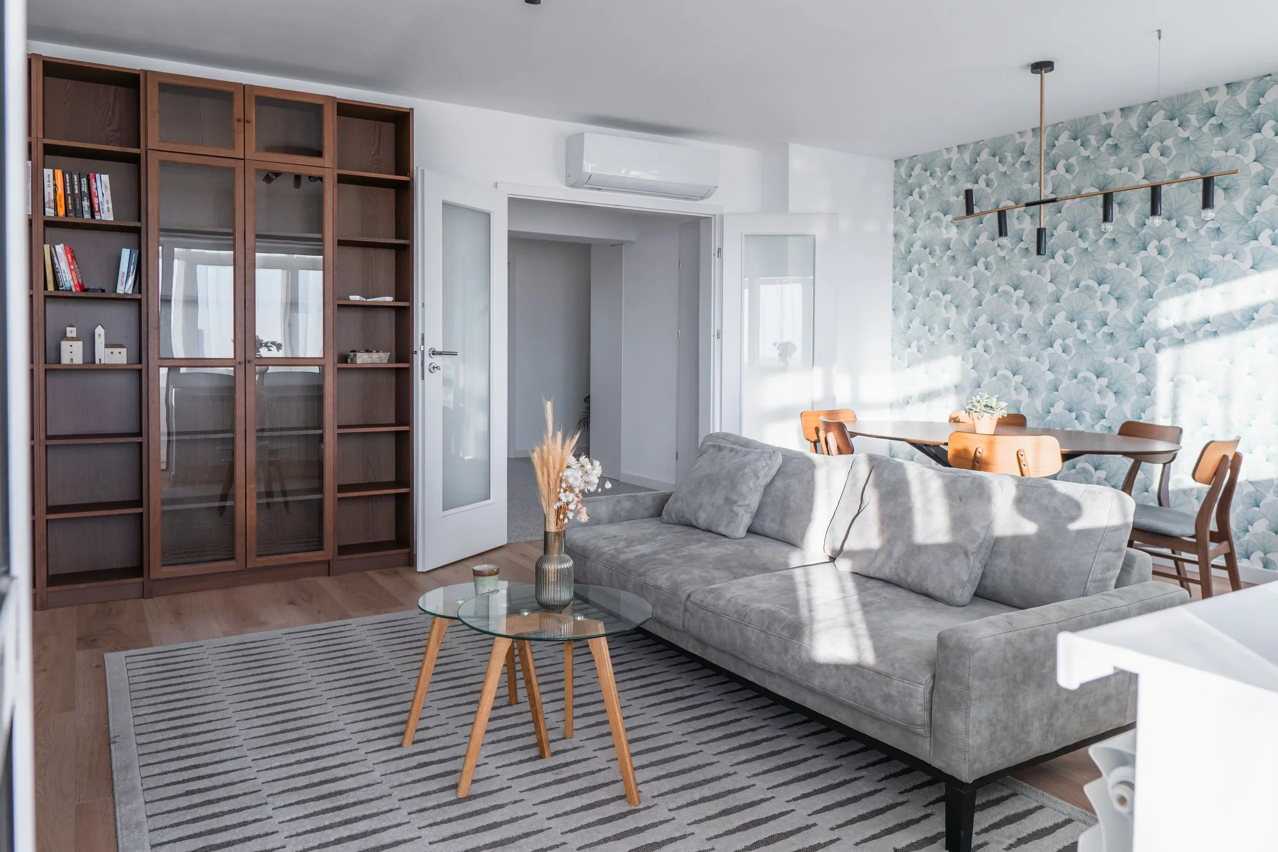 Bright living room with a gray sofa, glass coffee table, wooden bookshelf, and a dining area with a floral accent wall and wooden dining table with chairs.
