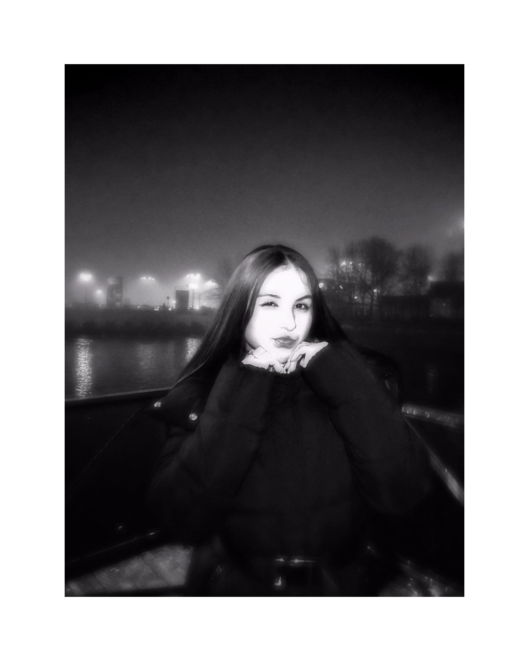 A young woman with long hair, wearing a dark jacket, winks and rests her chin on her hands, sitting outdoors near a body of water at night, with city lights and trees in the background, in black and white.