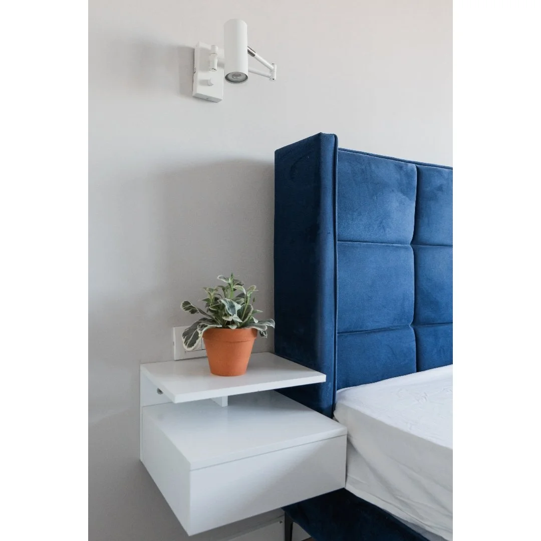 Close-up of a modern bedroom corner with a white nightstand holding a potted plant, a blue upholstered headboard, and a wall-mounted reading light on a white wall.