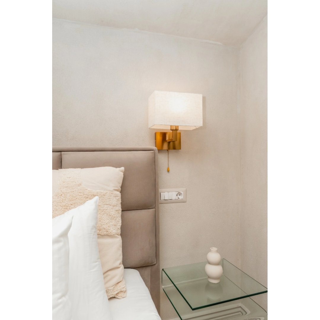 Wall-mounted lamp with white rectangular shade and gold base above a light grey upholstered headboard in a bedroom. Glass side table with a white vase next to the bed.
