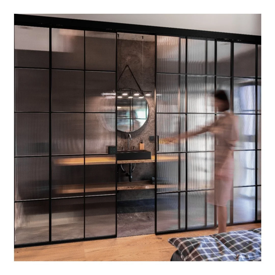 Interior of a modern bedroom with a glass sliding door separating the bathroom from the sleeping area. The bathroom has a concrete wall, a round mirror, a black sink, and a wooden shelf. A person dressed in a light-colored dress is pushing the sliding door open.