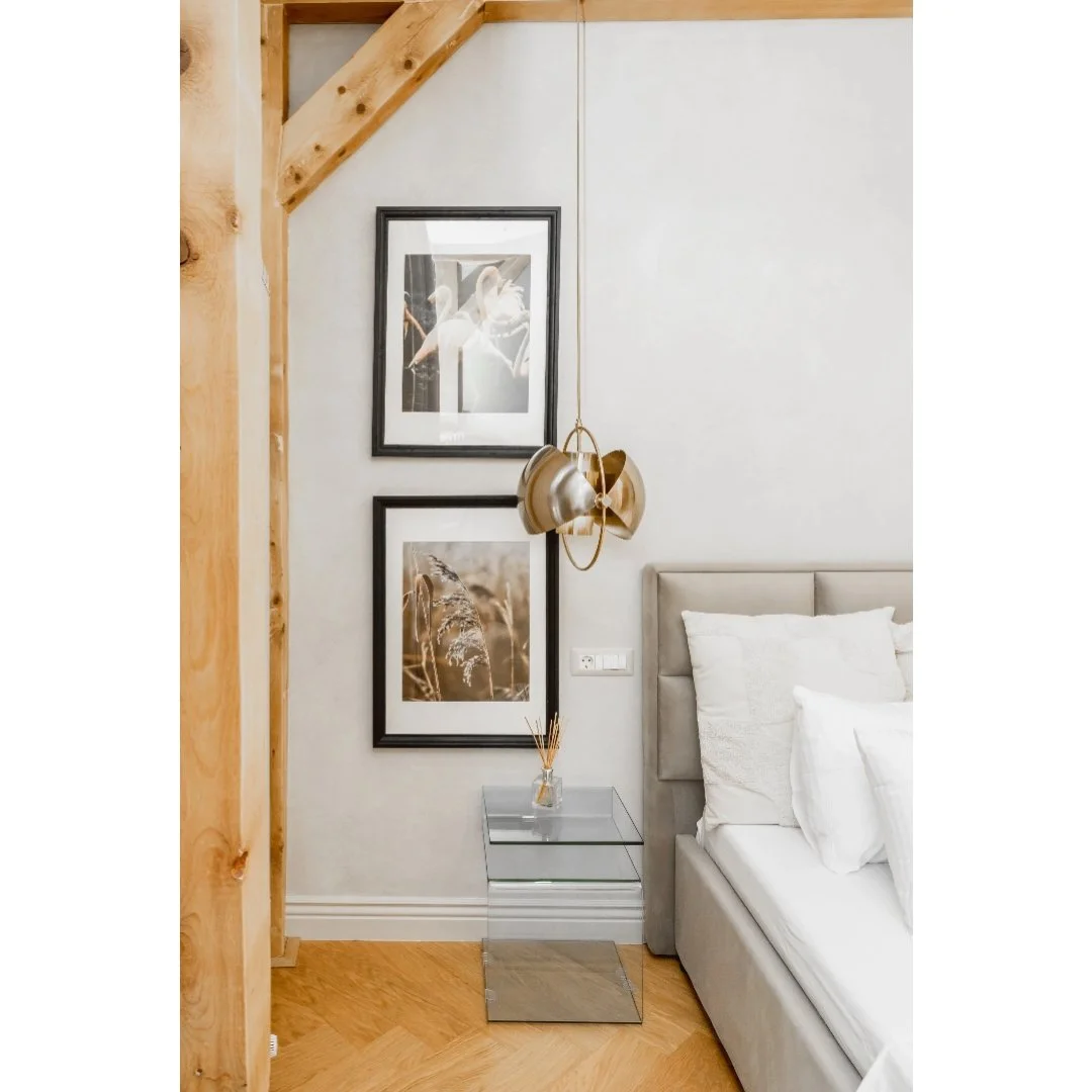 A bedroom corner with a gray bed, white pillows, a mirrored bedside table with a small vase, framed pictures of nature on a white wall, a modern gold hanging lamp, and wooden beams overhead.