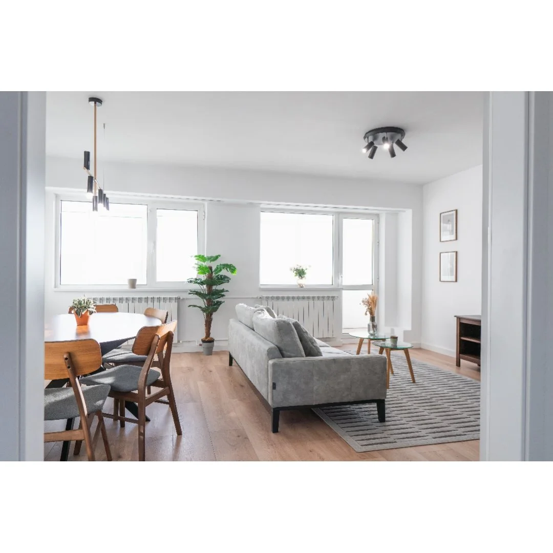 Bright living room with large windows, a gray sofa, and a wooden dining table with chairs. Decor includes potted plants, framed artwork, and a striped rug.