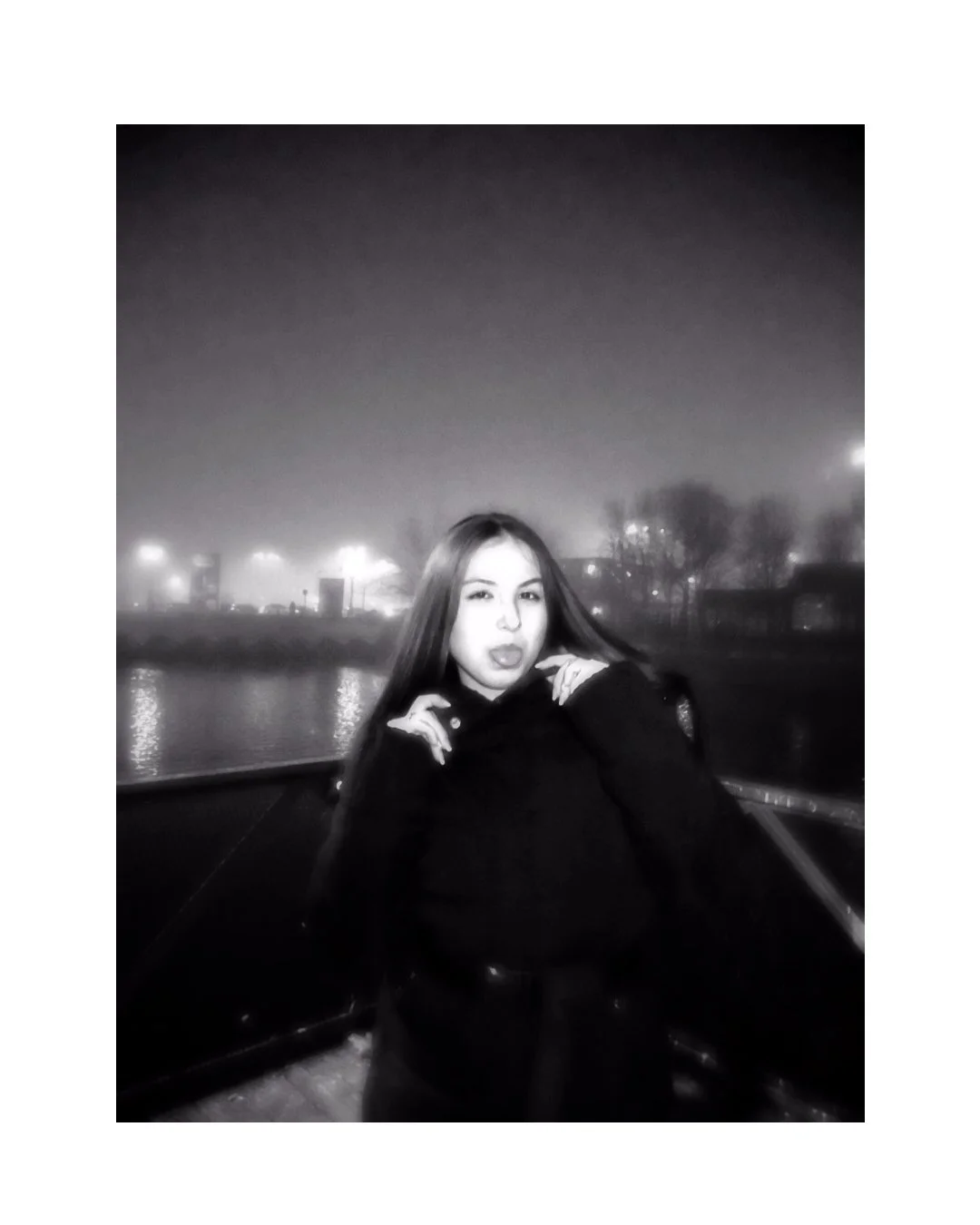 A young woman with long hair winks and poses with her hands near her face at night near a body of water, with city lights and trees in the background, in black and white.
