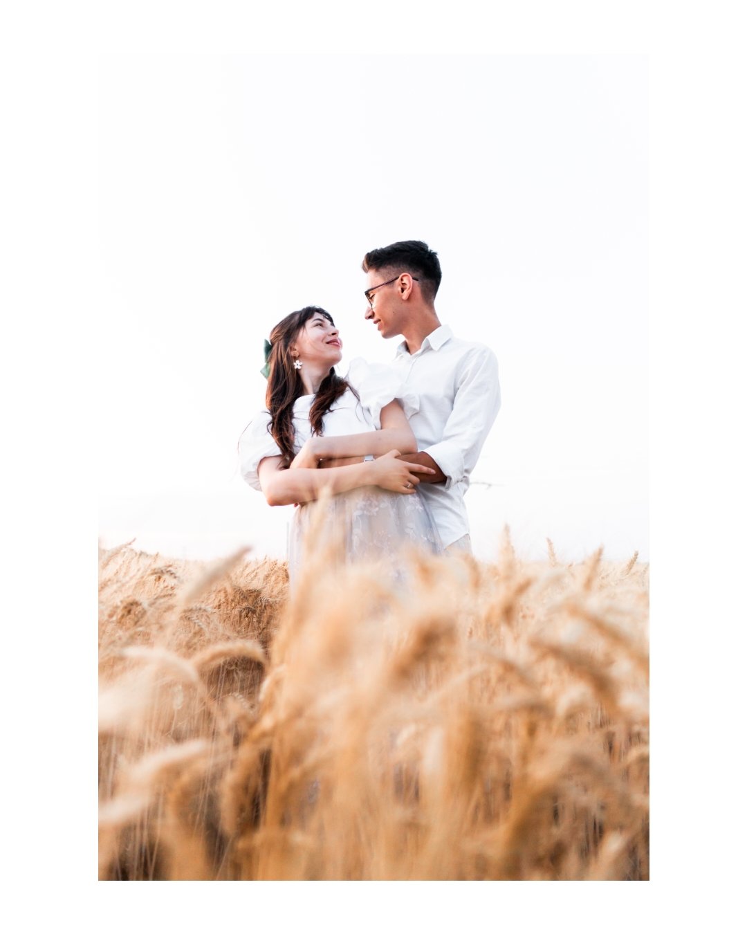 A young couple in white clothes standing in a wheat field, looking at each other affectionately.