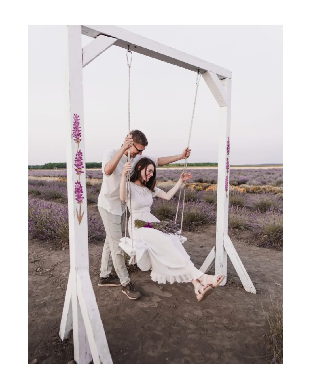 A couple on a white swing set in a lavender field, the woman sitting and holding a bouquet, the man pushing the swing, both smiling.