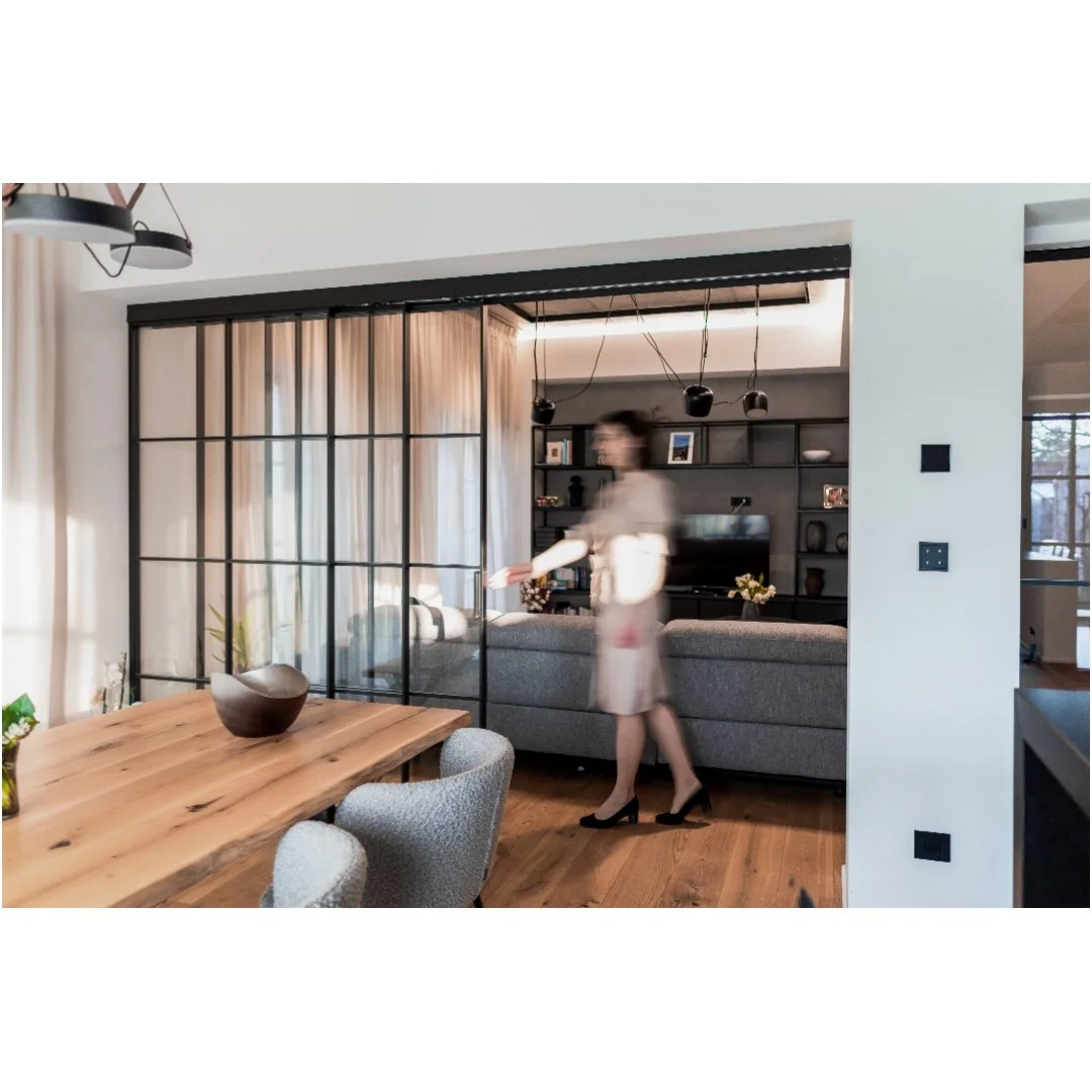 A woman in heels and a beige dress walking in a modern living room with a wooden dining table, gray chairs, a black glass sliding partition, a gray couch, wall shelves with decorative items, and a television.