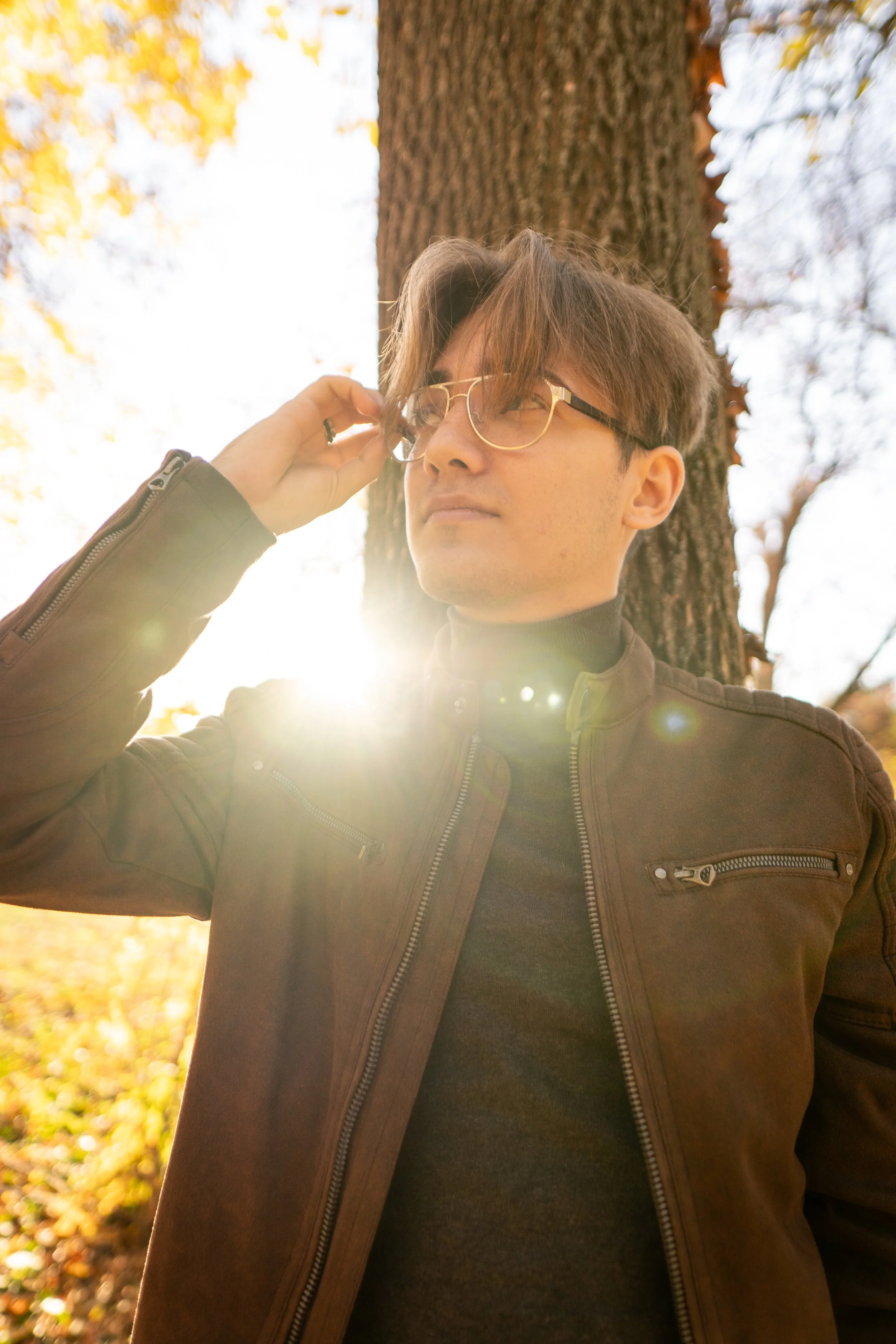 A man with brown hair and glasses standing outdoors near a tree, wearing a brown jacket, with sunlight shining from behind him.