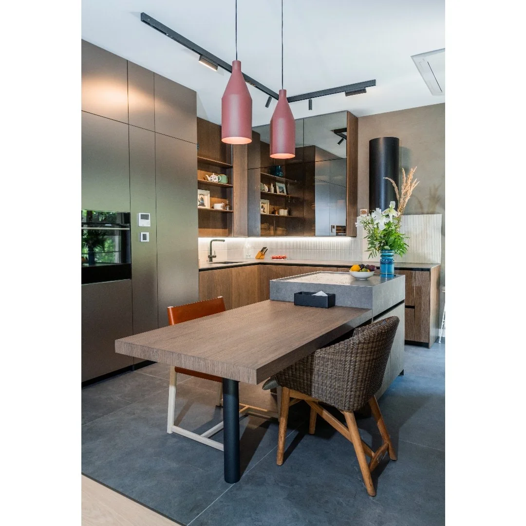 Modern kitchen with wooden cabinets, pink pendant lights, and a dining table with two chairs, one wicker and one wood, decorated with a vase of flowers and a bowl of fruit.