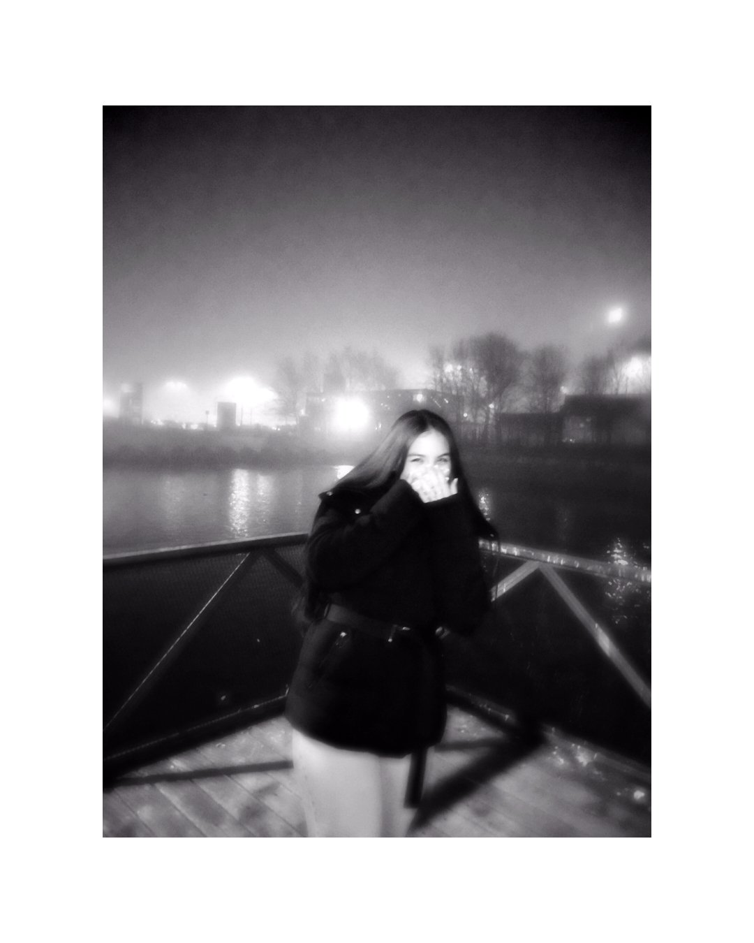 A woman standing on a bridge at night, covering her mouth and nose with her hand, with mist or fog in the background and city lights reflecting on the water.