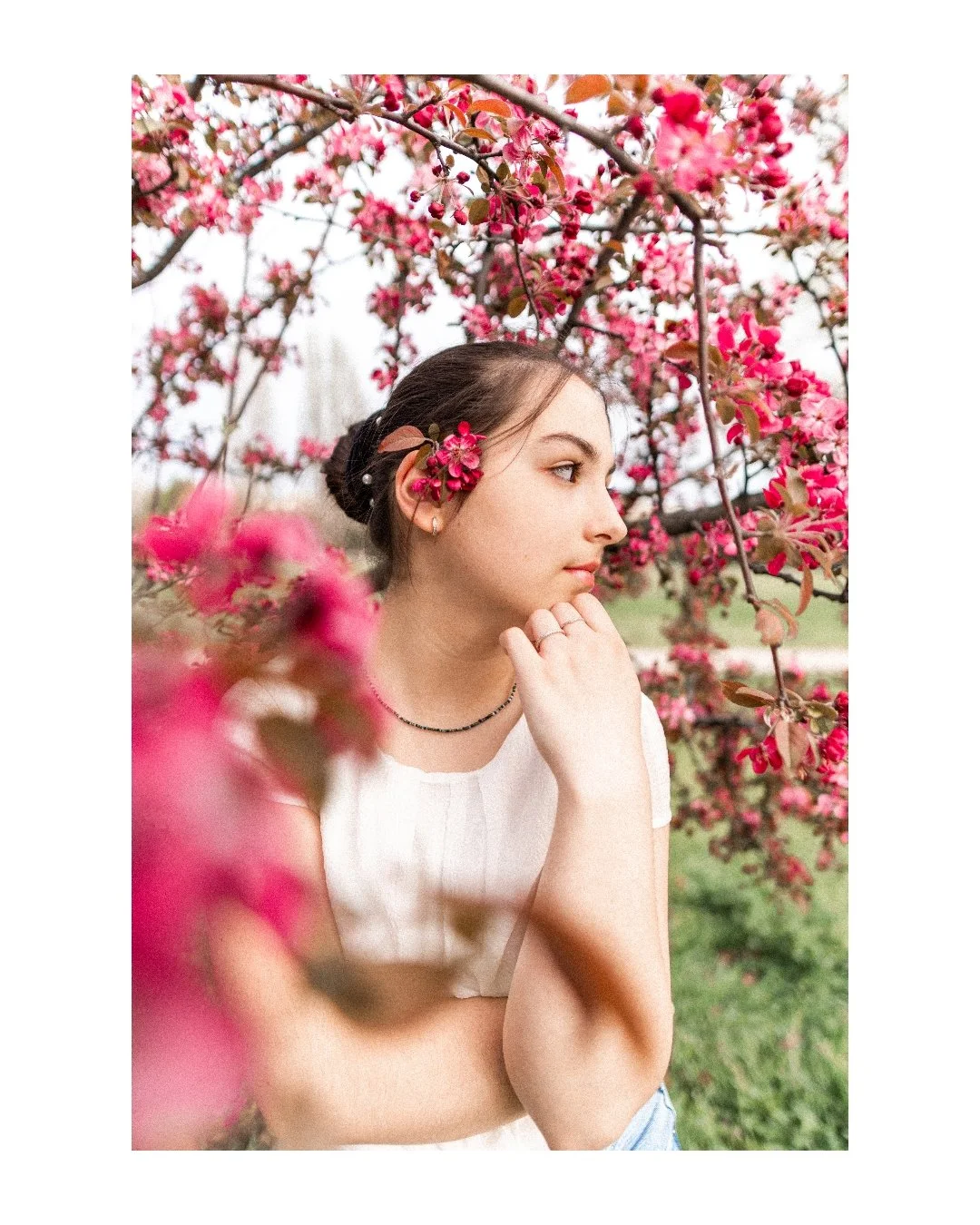 Young woman with dark hair decorated with pink flowers, standing among blooming pink cherry blossom trees, wearing a white top, in a serene outdoor setting.