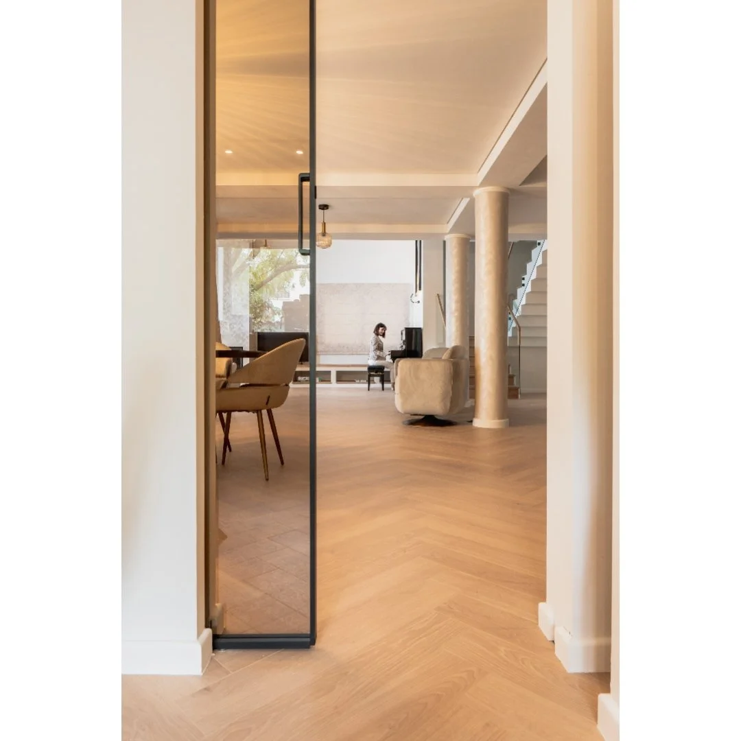 Living room with a woman playing piano, beige chairs, and a staircase, viewed through a black glass door.