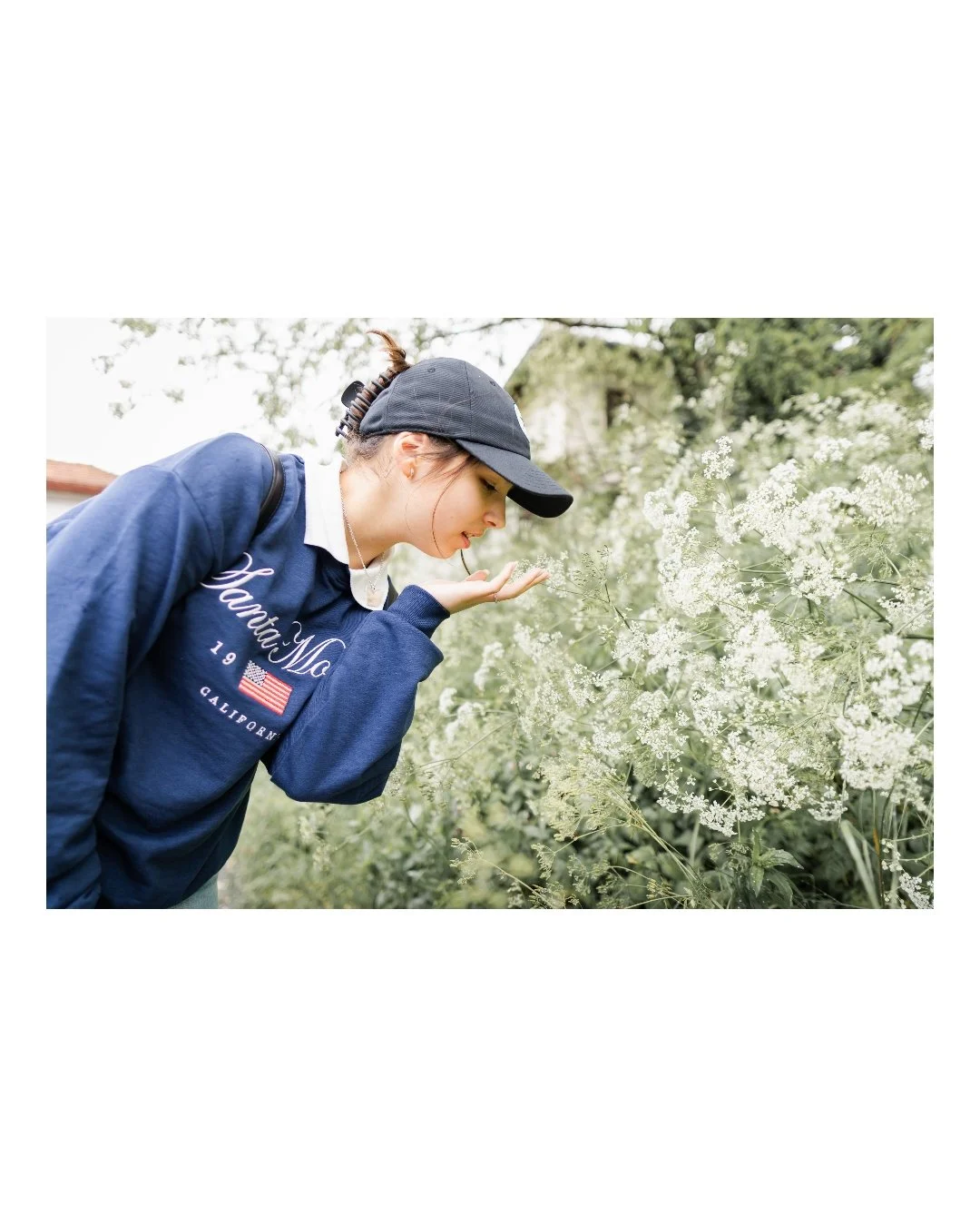 Young woman wearing a dark baseball cap and a blue sweatshirt smelling white flowers in a garden.