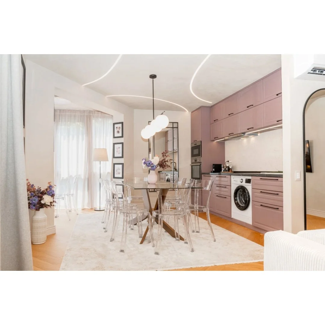 Open-concept kitchen and dining area with pink cabinetry, glass dining table, transparent chairs, and decorative lighting fixtures.