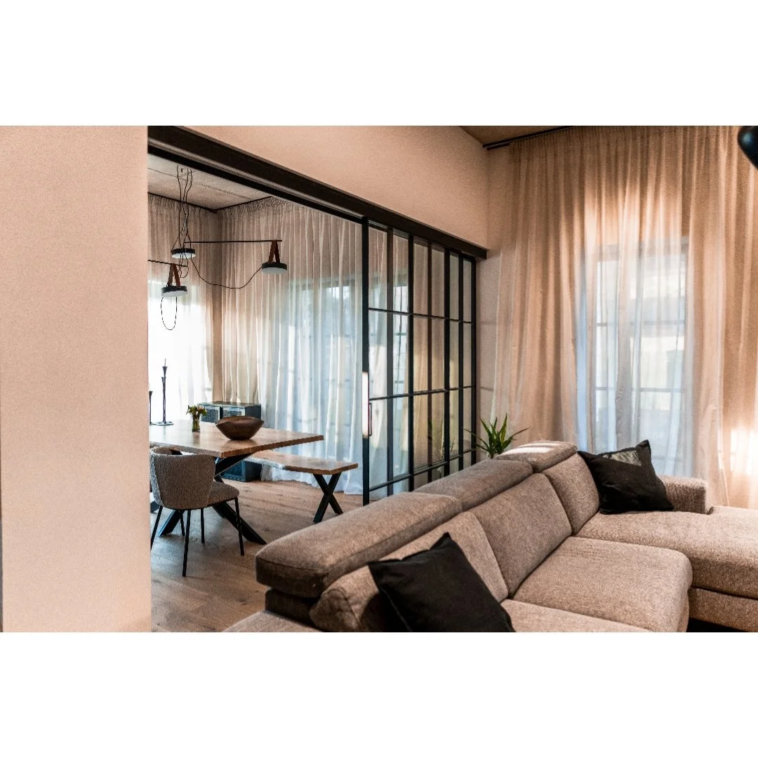 Living room with a beige sectional sofa, black pillows, sheer curtains, and a glass sliding door separating to a dining area with a wooden table, chairs, and window with curtains.
