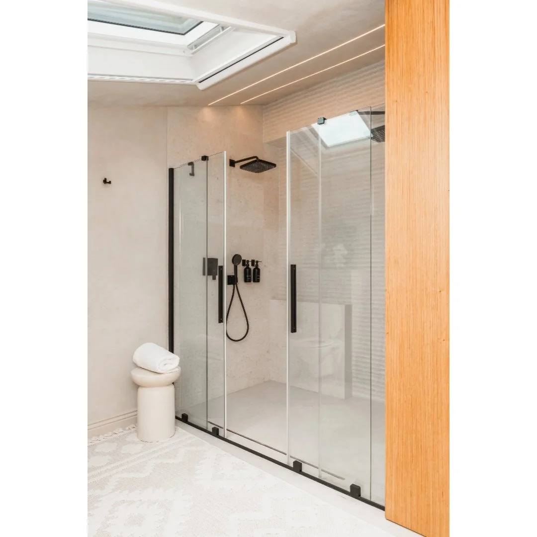 Modern walk-in shower with glass doors, black fixtures, and a wooden wall panel, located in a bathroom with a light-colored wall and ceiling, a skylight, and a rolled towel on a stool.