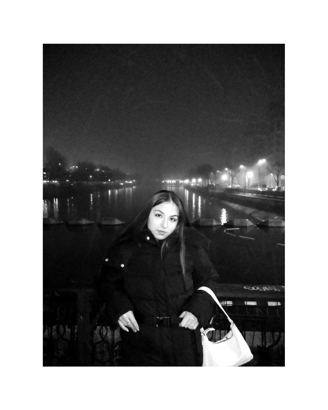 A young woman standing outdoors at night, wearing a dark jacket and holding a white bag, with a river and streetlights in the background, captured in black and white.