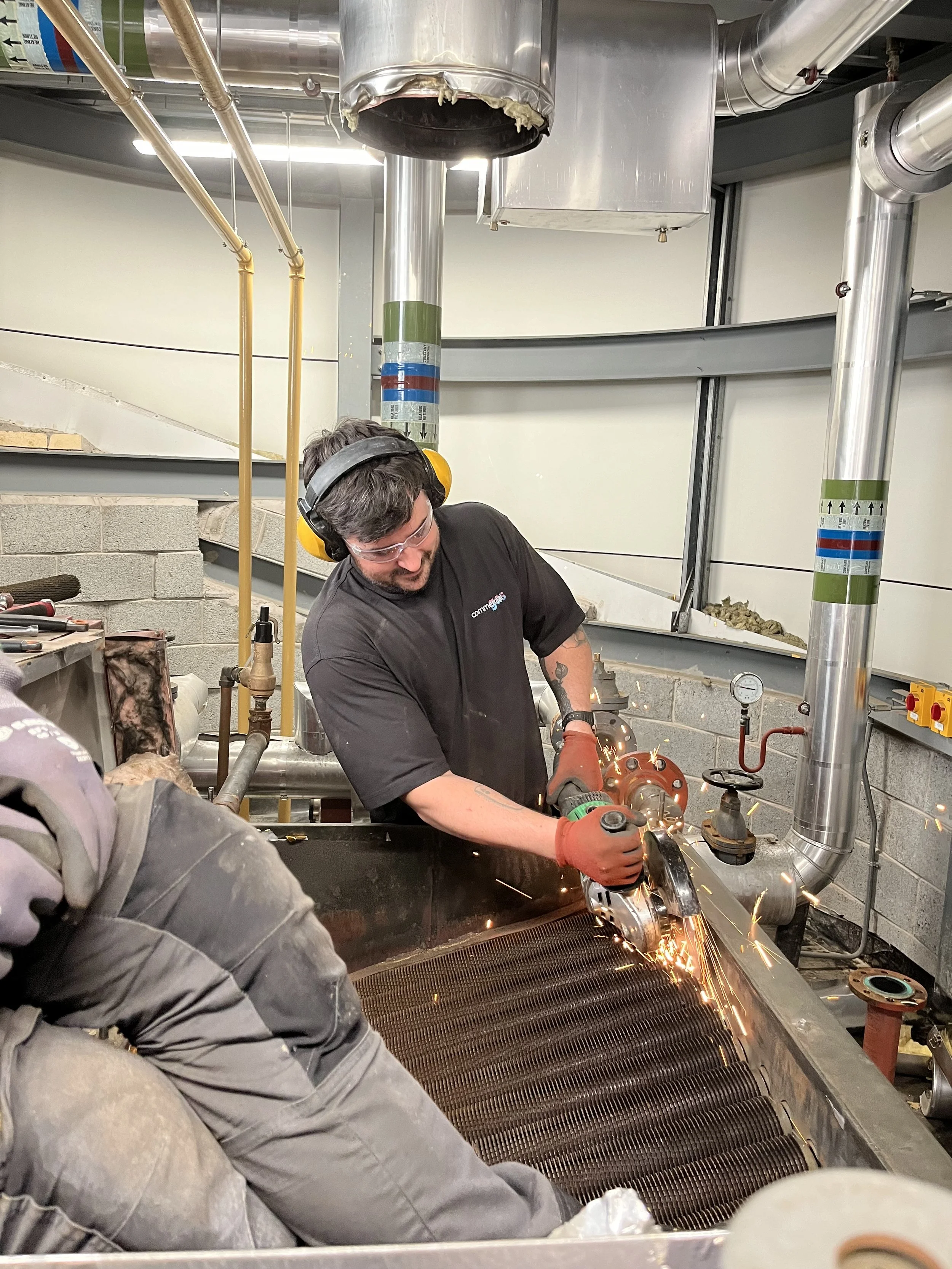 Man wearing safety goggles, headphones, and gloves using a power tool to cut or grind a large metal structure, sparking debris.