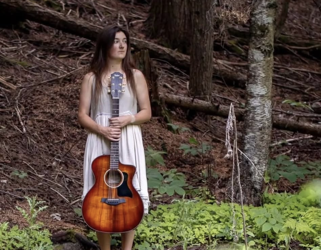 Young woman musician holding a guitar in the woods.