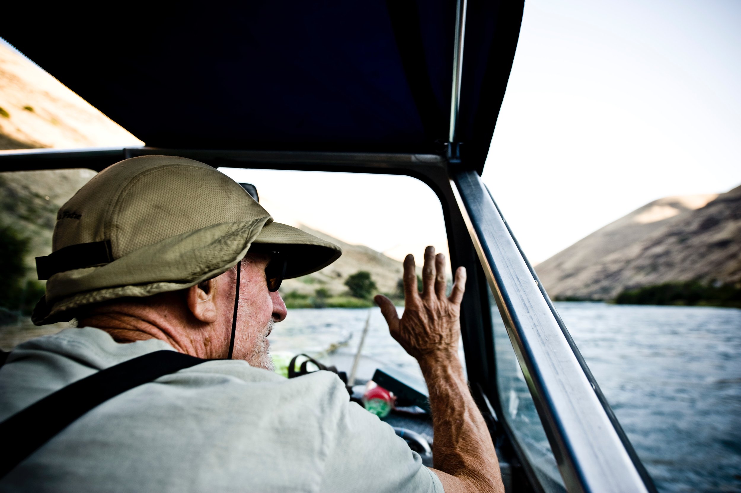 An elderly man wearing a beige hat and sunglasses, steering a boat on a river with mountains in the background.