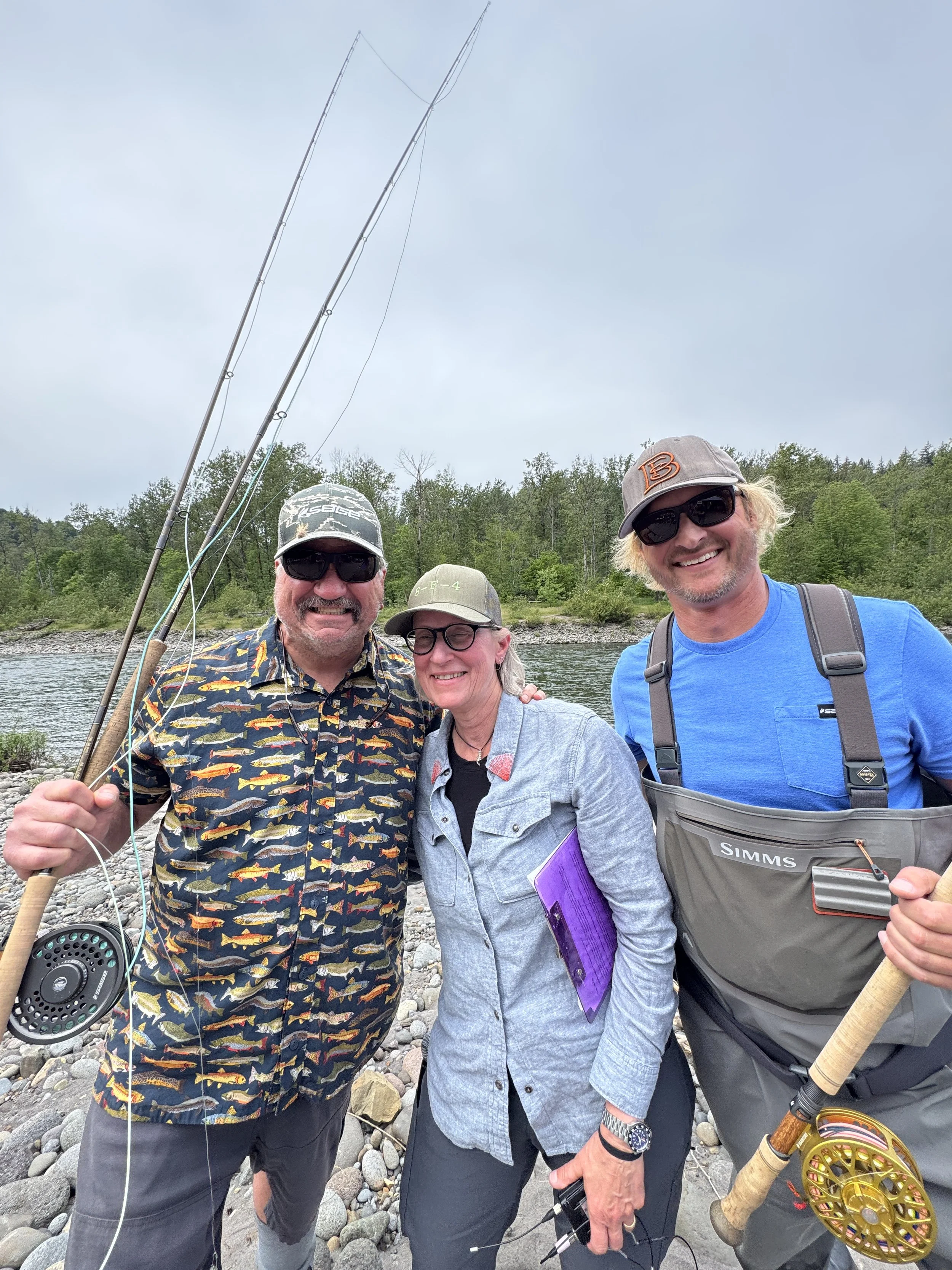 Three people smiling and posing on a rocky riverbank, holding fishing rods, with trees and a cloudy sky in the background.