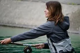 Woman fishing in a body of water, wearing a dark jacket and carrying a backpack.