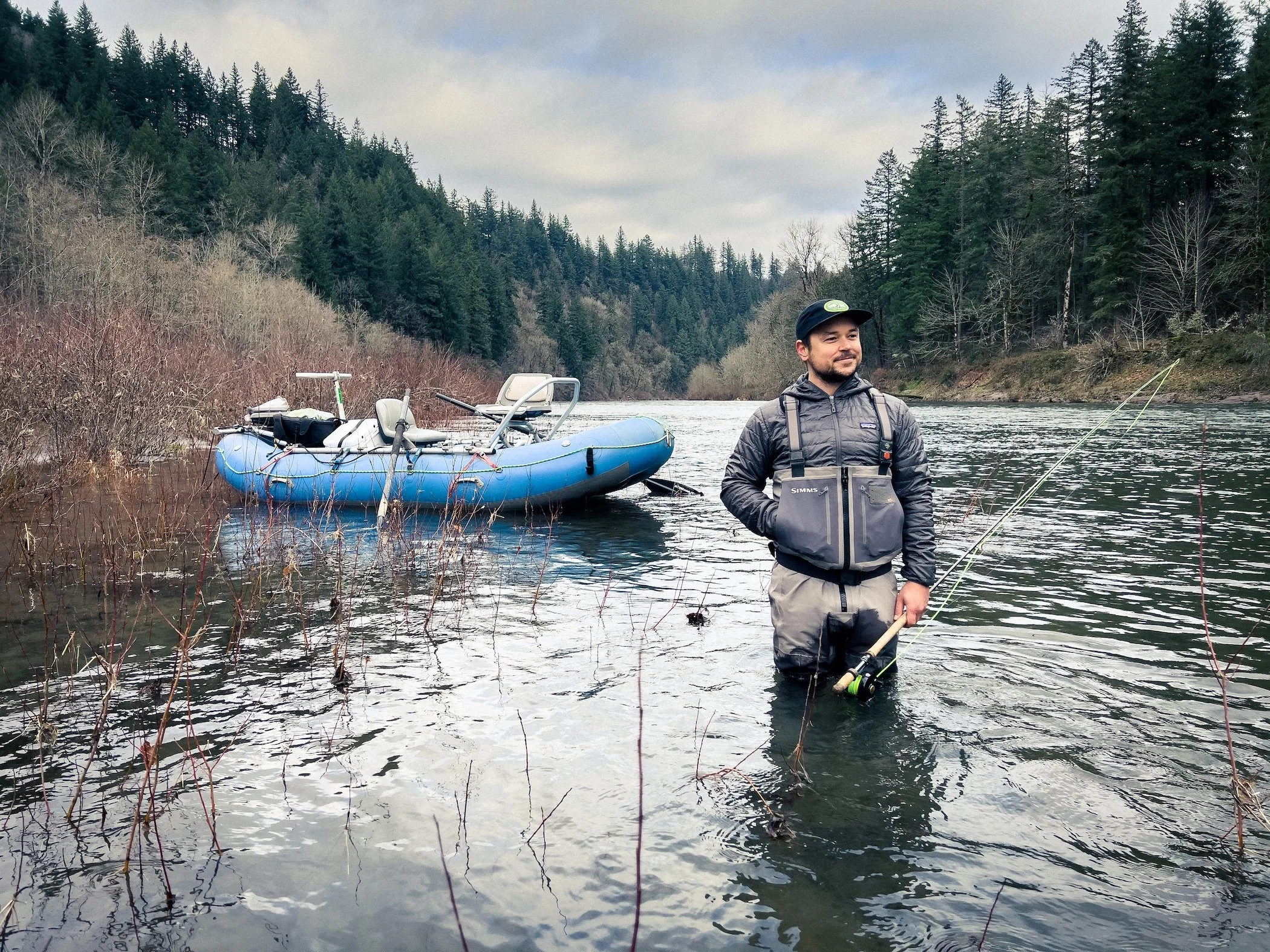 A man in fishing gear standing in a river with a fishing rod, with a blue inflatable boat on the riverbank beside him, surrounded by trees and forested hills on a cloudy day.