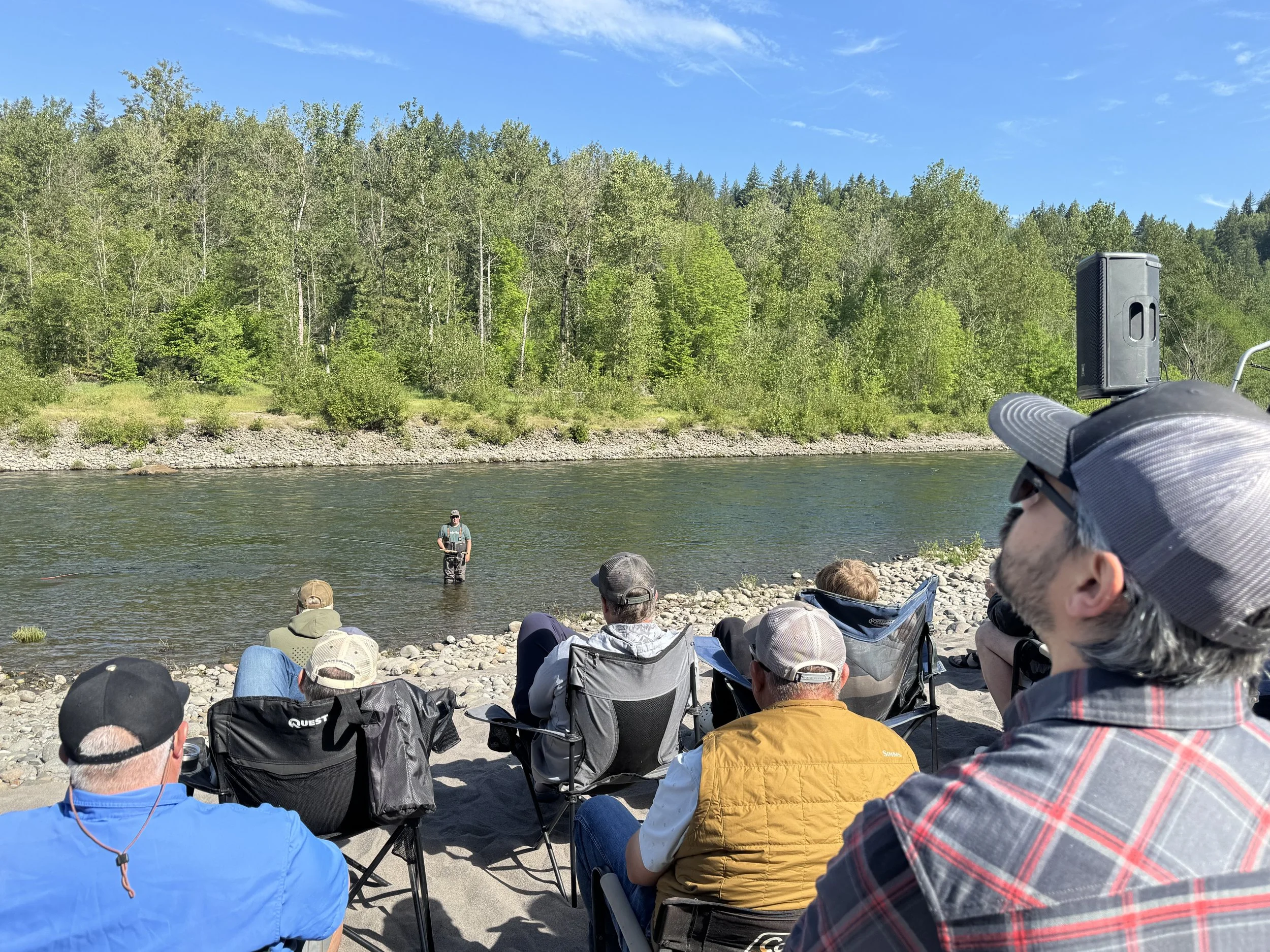 People sitting in chairs on a beach beside a river, watching a man fly fishing in the water with a forest and blue sky in the background.