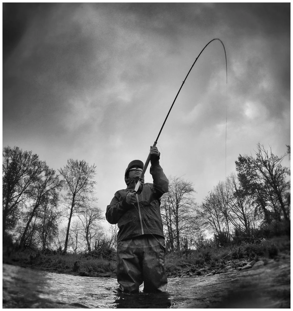 A person standing in a stream fishing with a fishing rod, surrounded by trees under a cloudy sky.