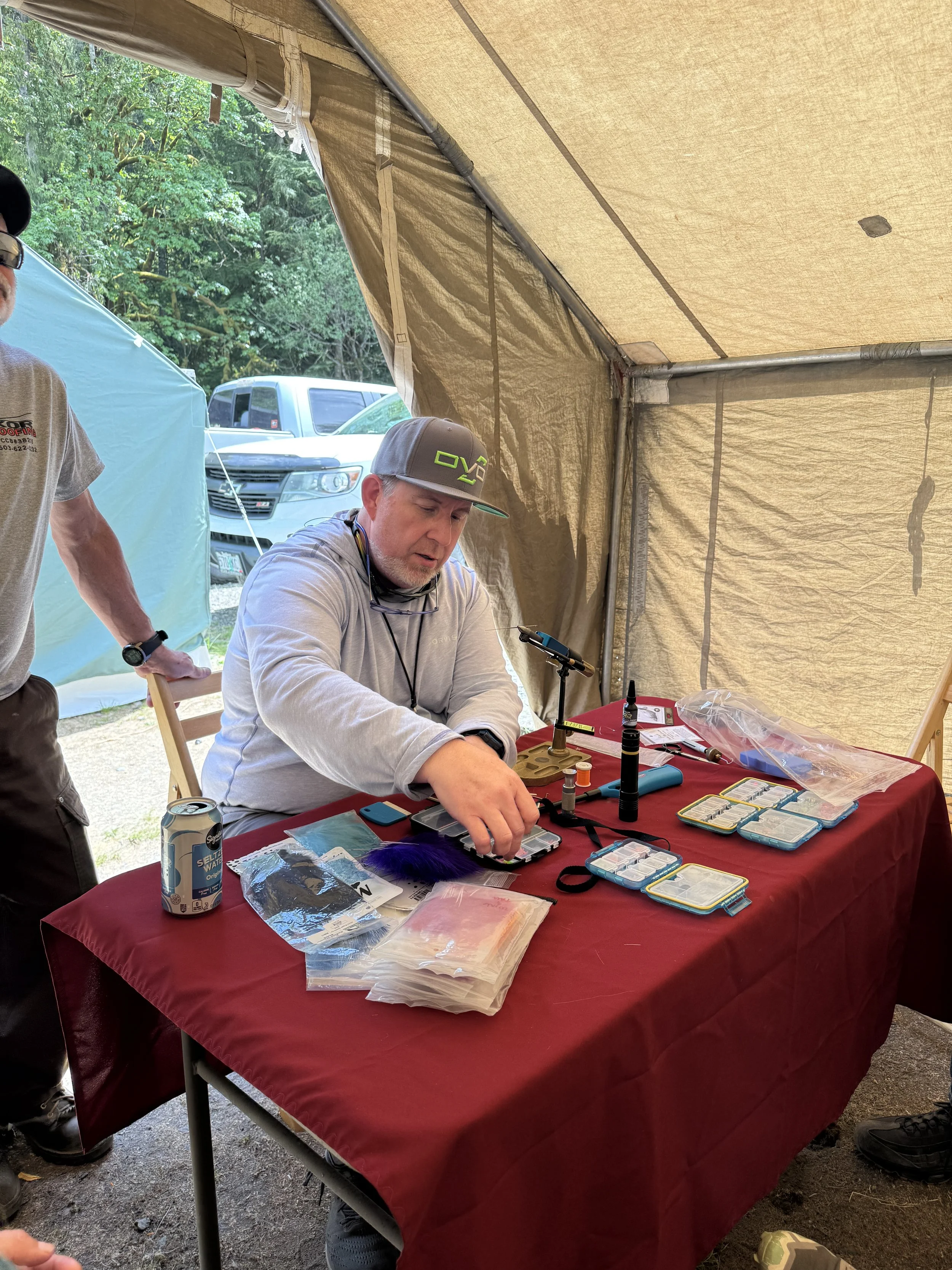 A man sitting at a red table assembling a fly fishing lure inside a beige tent with another man partially visible to the left. Various small containers and tools are on the table.