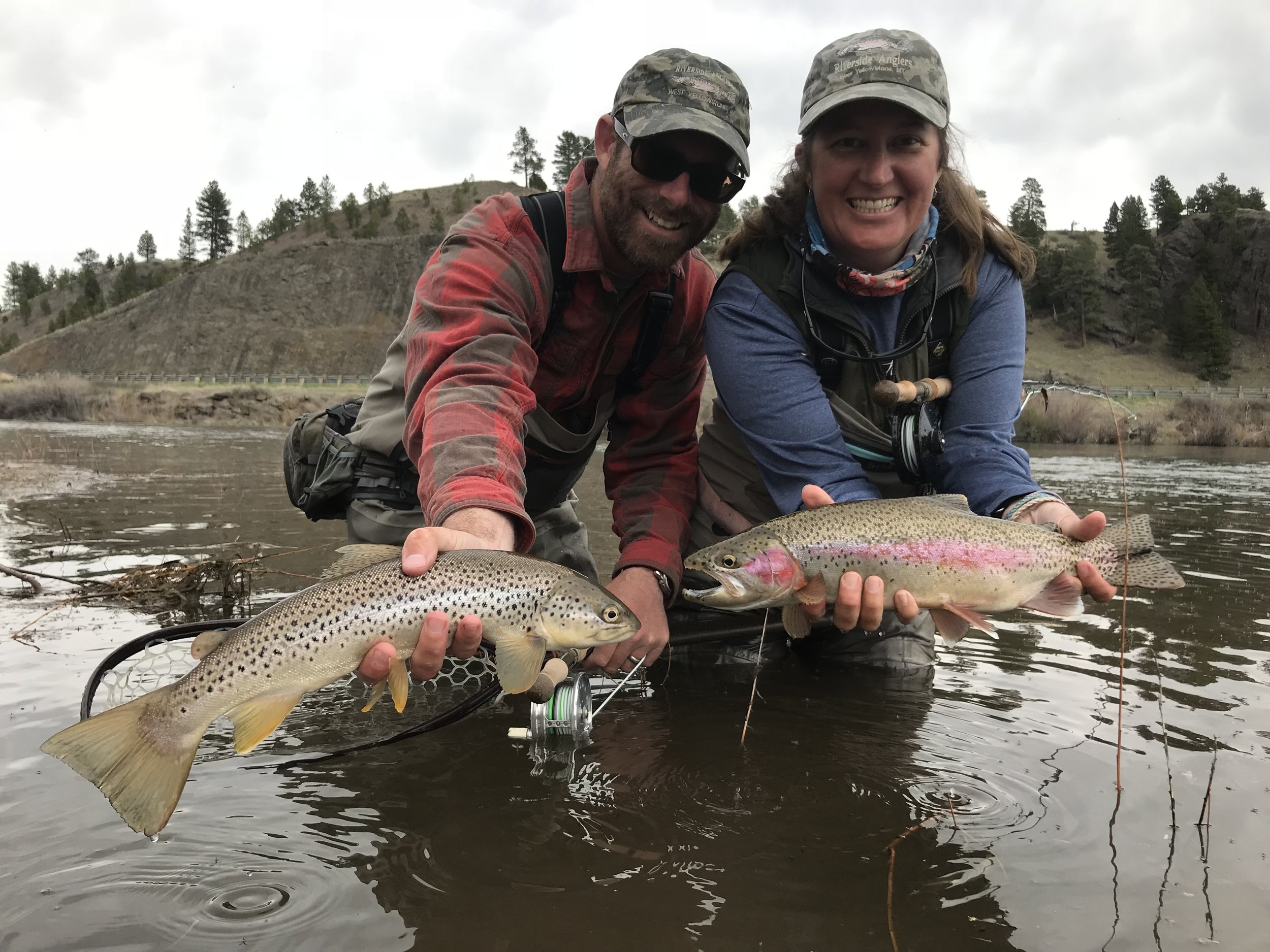 A man and woman standing in a river, each holding a large fish they caught while fishing, with hills and trees in the background.