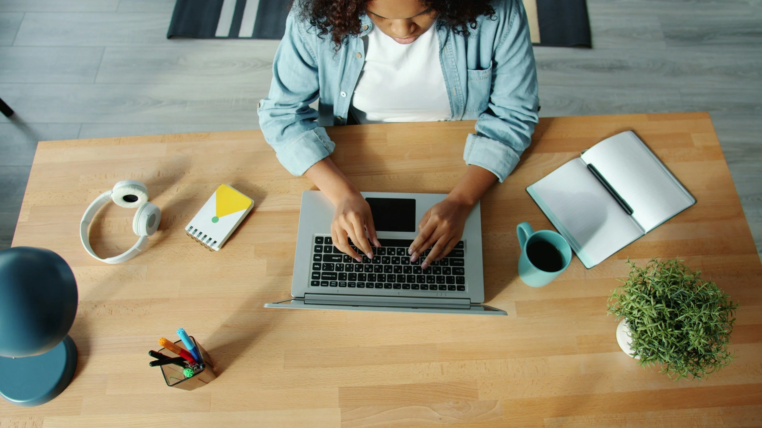 Top-down view of a woman working on a laptop at a wooden desk. The desk has a pair of headphones, a yellow and white notepad, a cup of coffee, an open notebook with a pen, a small potted plant, and a container holding colorful pens. The woman is wearing a light denim shirt and a white t-shirt.
