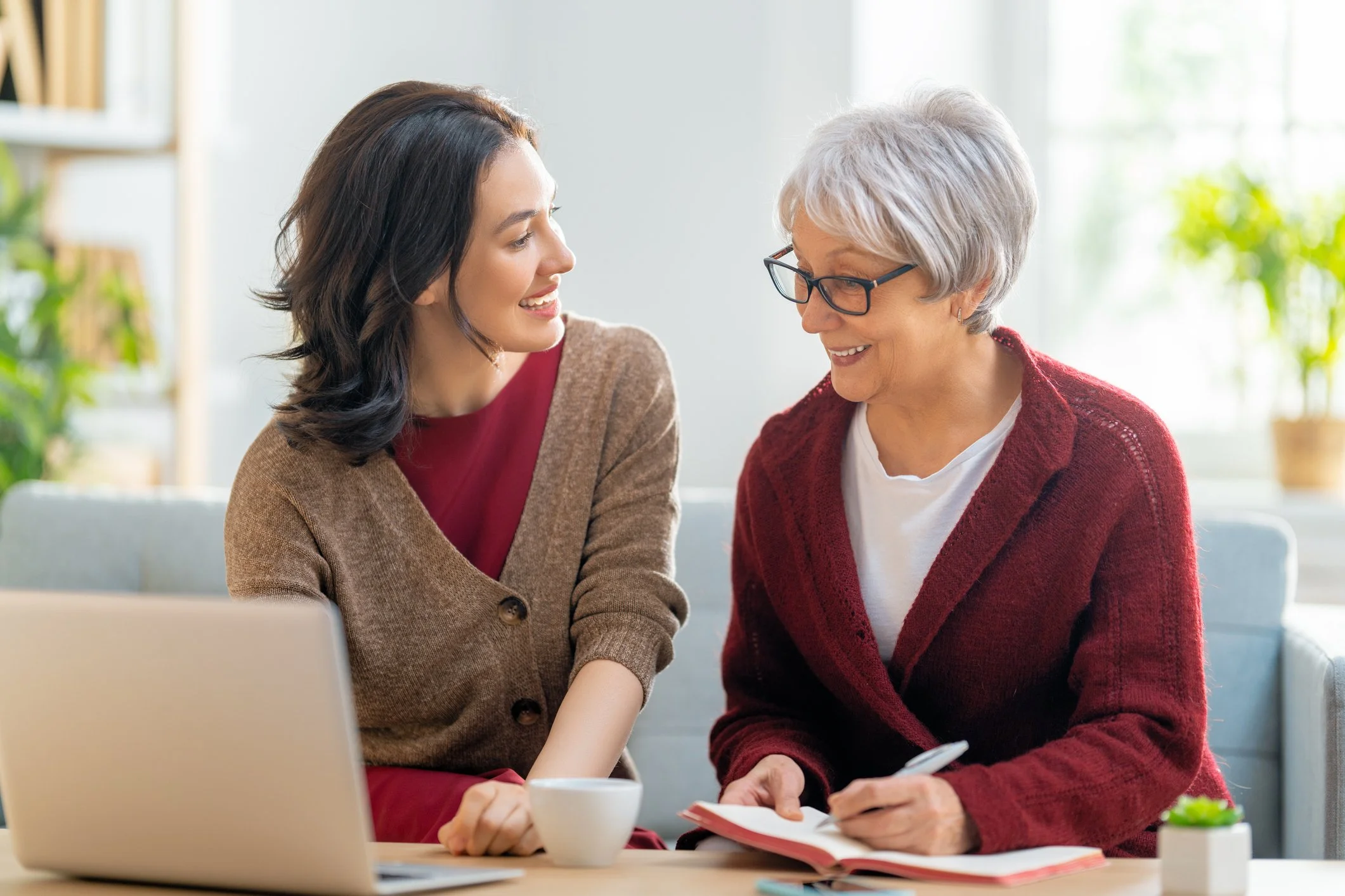 A young woman and an older woman are having a conversation at a table, both smiling, with a laptop, a cup, and a notebook on the table.