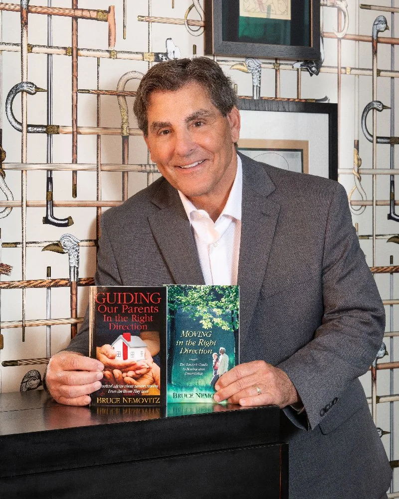 A man in a gray suit smiling at the camera, sitting behind a black table, holding two books titled "Guiding Our Parents in the Right Direction" and "Moving in the Right Direction." The background features a decorative wall with various saws and tools arranged on it.