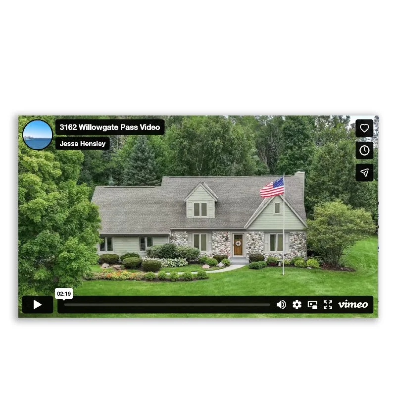A house with gray walls, stone accents, and a gabled roof, situated in a lush green yard with trees and landscaped bushes, featuring an American flag on a flagpole in front.