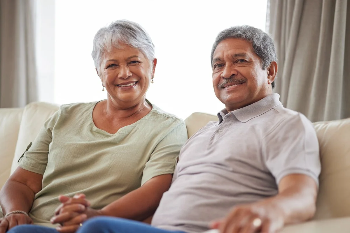 An elderly couple sitting on a cream-colored couch in a living room, smiling at the camera with sunlight streaming through the windows behind them.