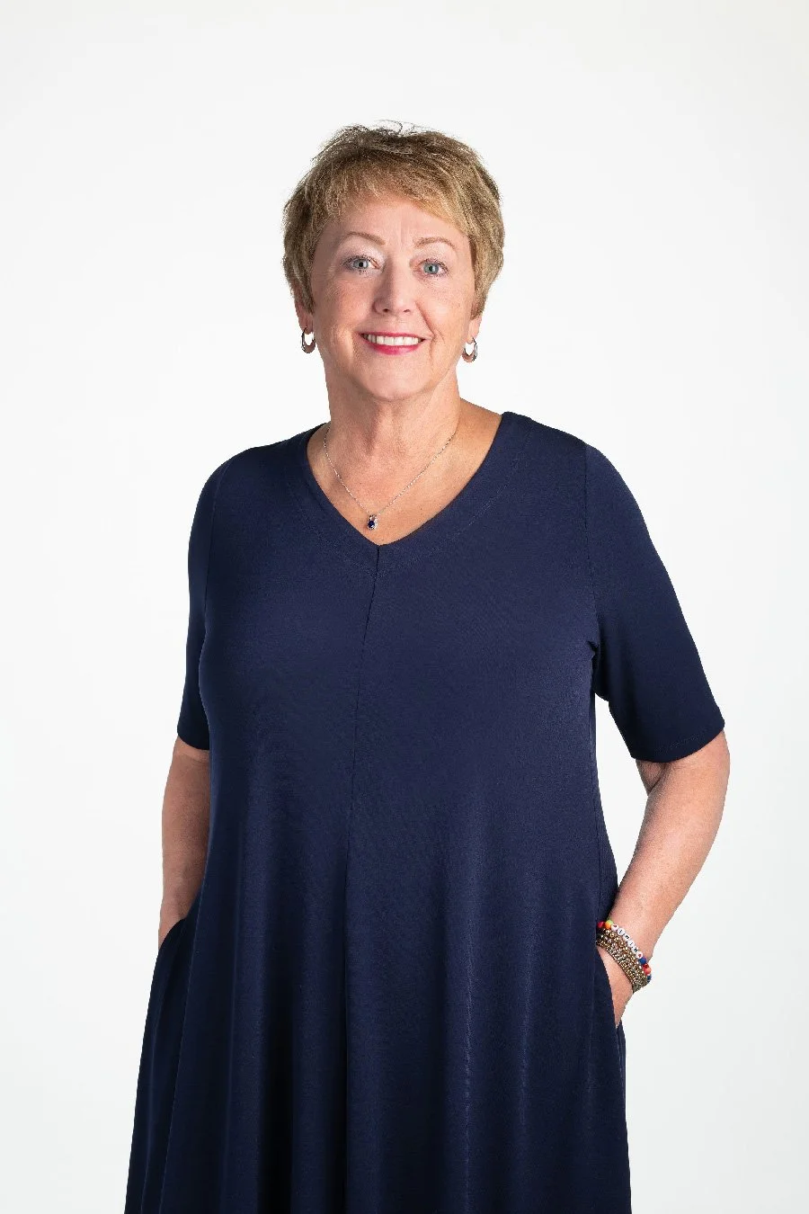 A middle-aged woman, Cindy, with short blonde hair, wearing a navy blue dress and jewelry, standing against a plain white background, smiling at the camera.
