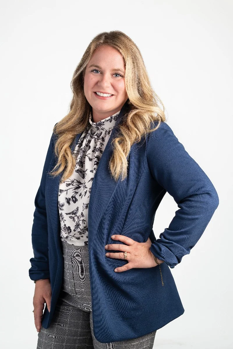 A woman, Erica Petri,  with long blonde hair wearing a navy blue blazer, a white blouse with black floral print, and gray plaid pants. She is smiling and posing with her right hand on her hip.