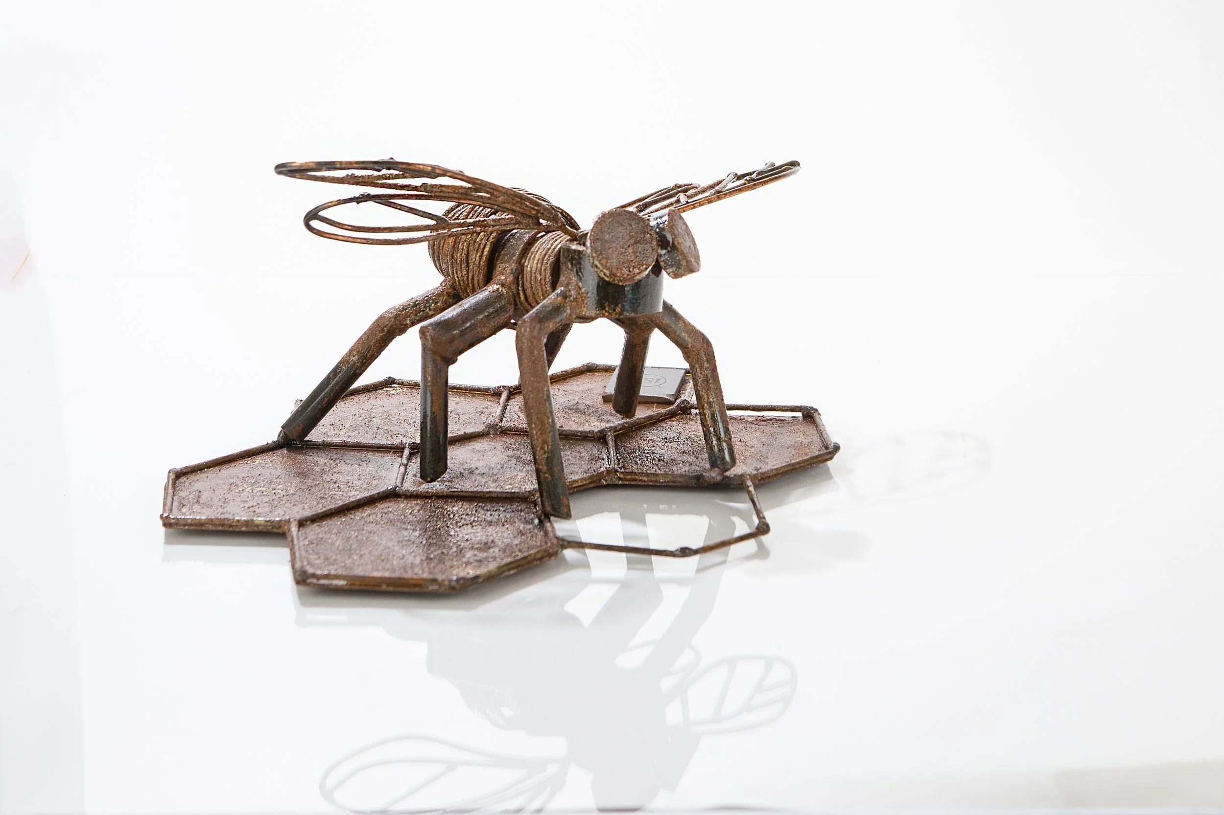Rusty metal sculpture of a bee on a hexagonal honeycomb-shaped base, reflected on a glossy surface.