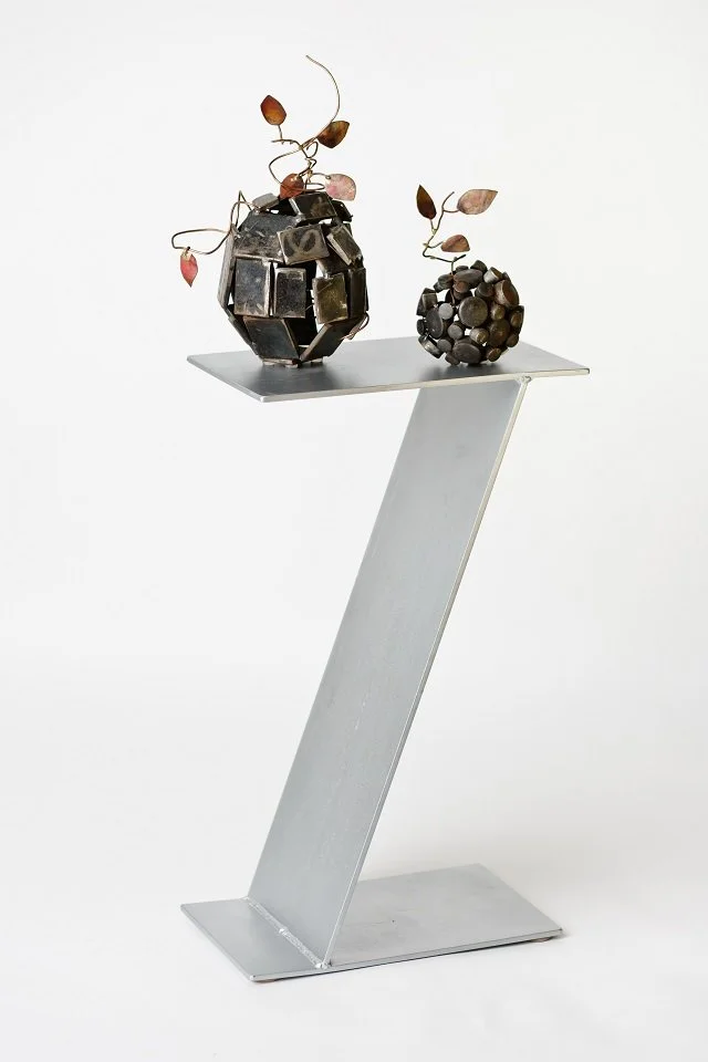Two decorative metal sculptures resembling abstract vases with small metal leaves on curly stems, displayed on a modern white pedestal against a white background.