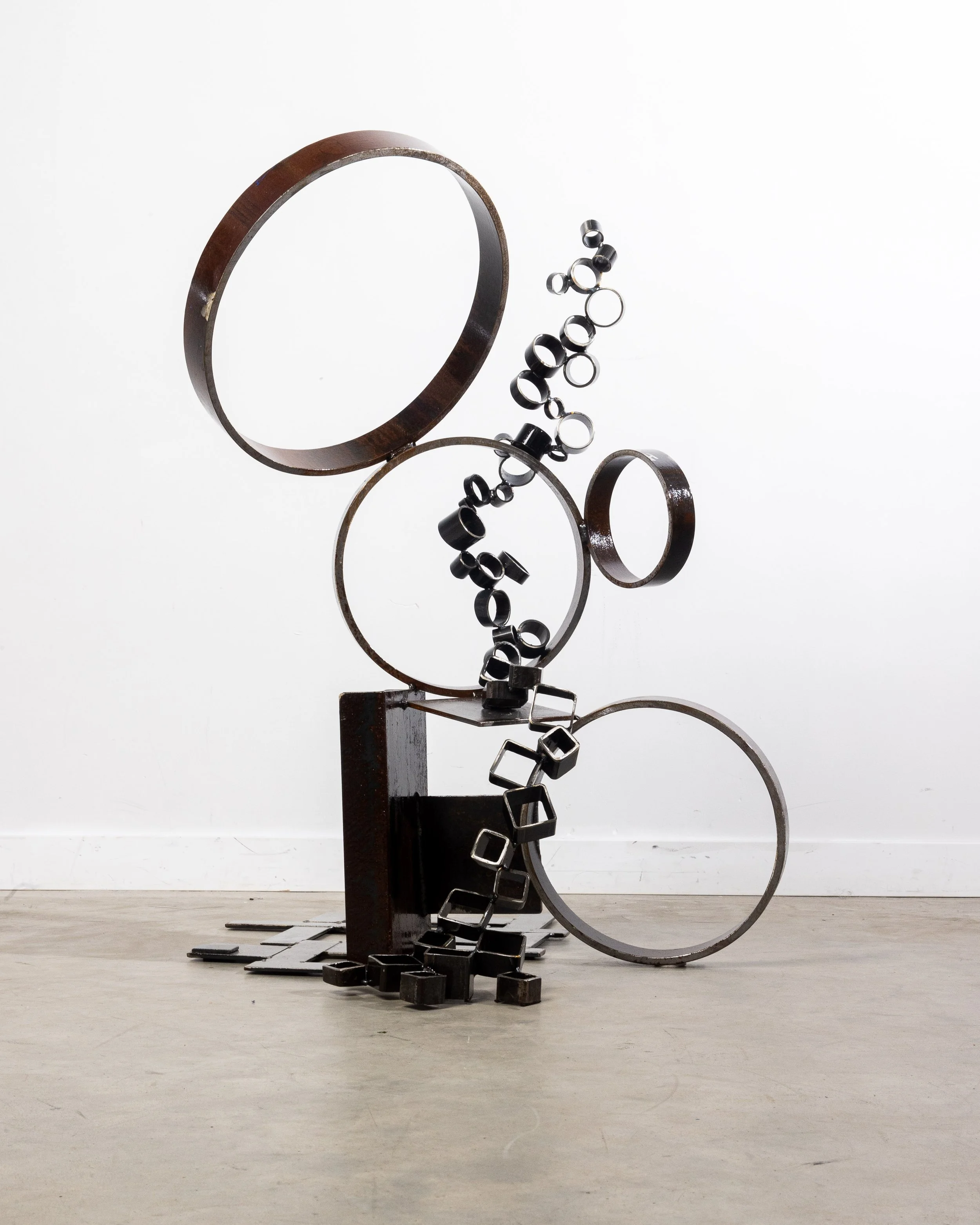 Abstract sculpture made of black metal rings and squares arranged on a wooden base against a white wall.