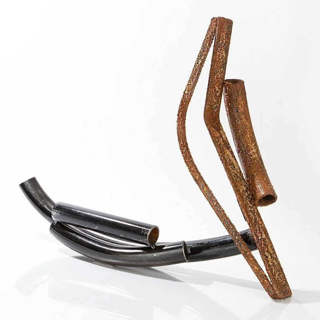 An abstract sculpture made from black metal pipes and rusted metal rods against a white background.