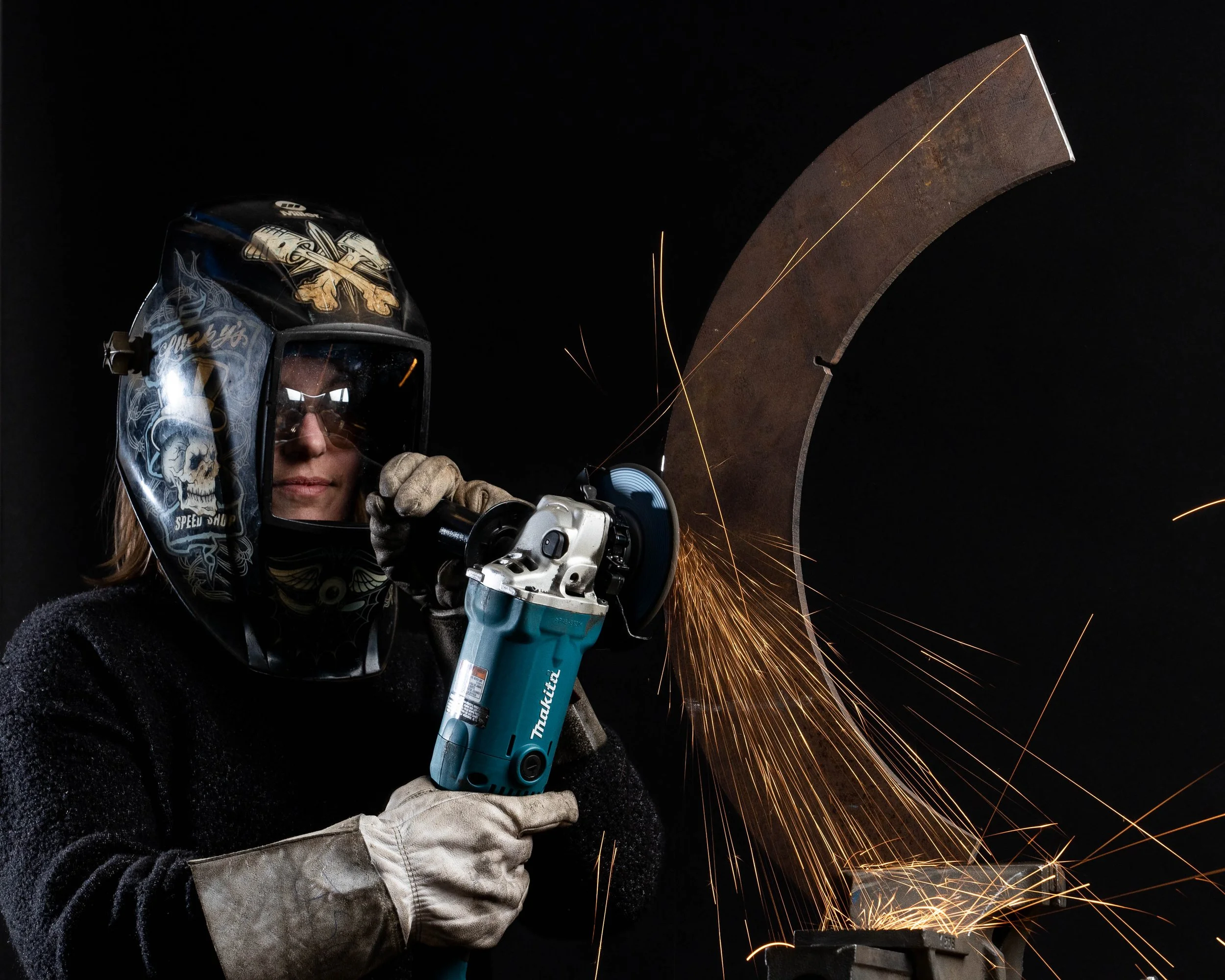 A woman wearing a face shield, goggles, gloves, and a black sweater is using a cutting tool to cut a large metal piece, creating sparks against a black background.