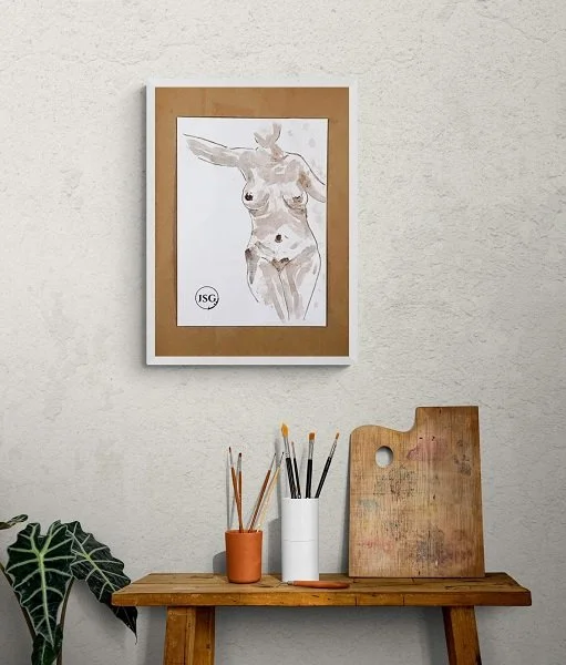 A framed watercolor painting of a deer on a beige wall above a wooden table with art supplies, including brushes and a wooden palette, and a green plant nearby.