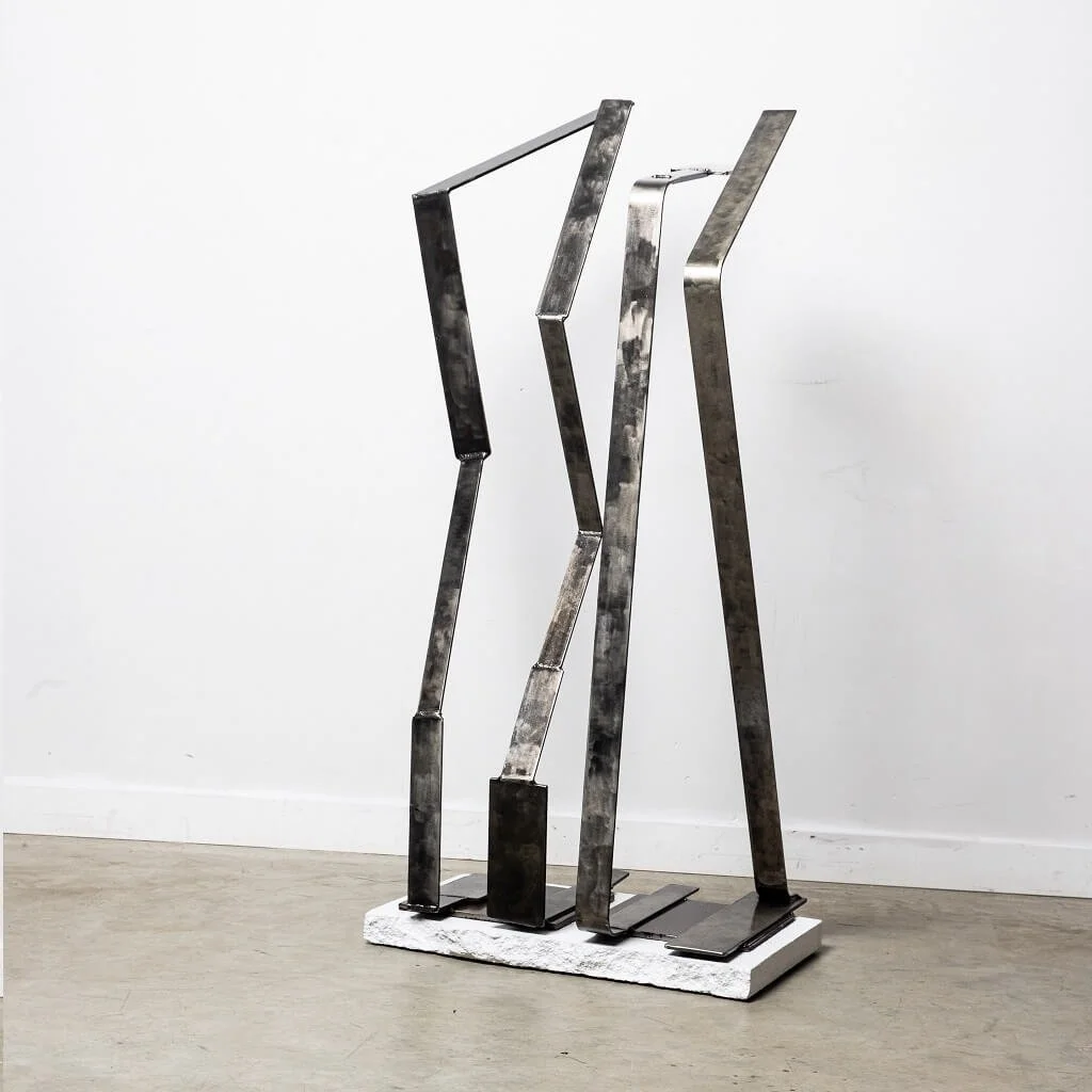 Abstract sculpture made of metallic irregularly shaped panels on a white base, placed against a plain white wall.