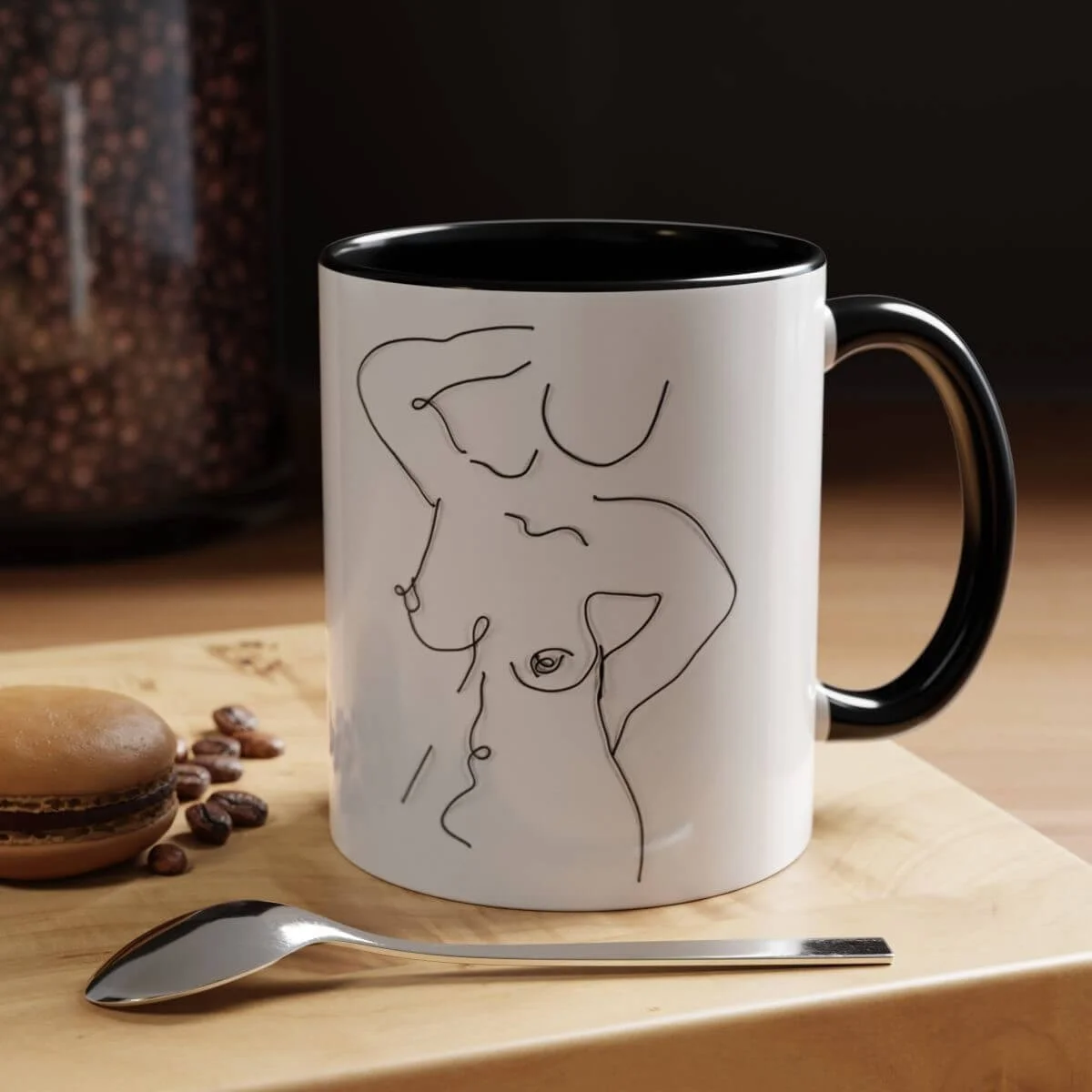 A white mug with a black interior and handle, featuring a minimalist black line drawing of a nude woman with one arm behind her head. The mug is on a wooden surface next to coffee beans, a spoon, and a macaron. A jar of coffee beans is visible in the background.