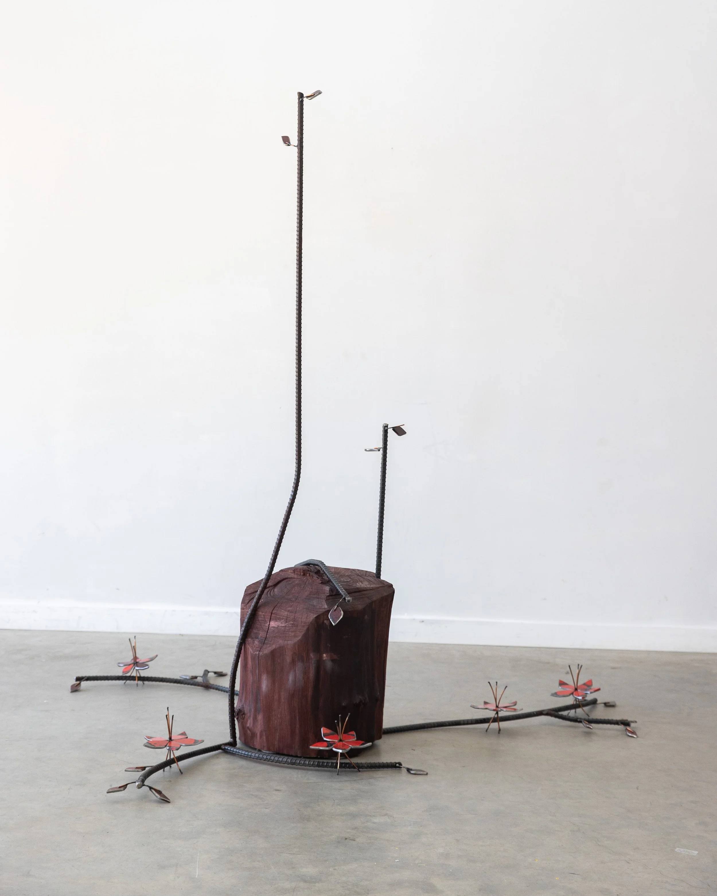 An art installation featuring a large wooden block with various metal and wooden elements, including tall metal rods with attached lights, metal flowers on the ground, and a metal insect sculpture, all set against a plain white wall and gray floor.