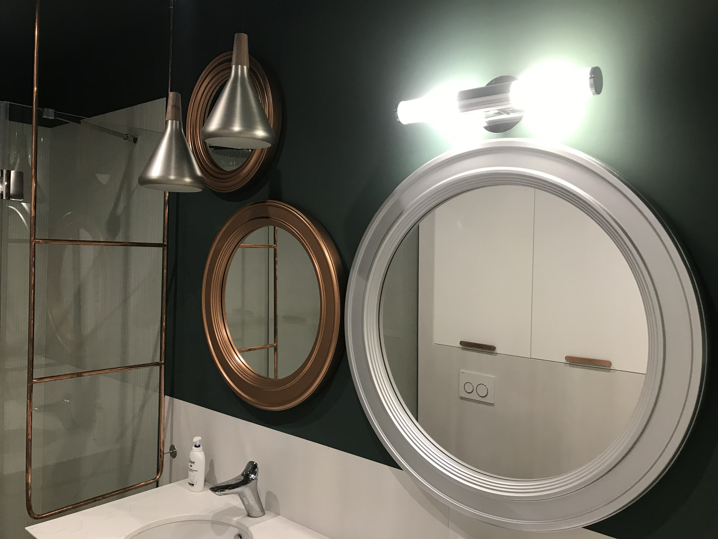 Bathroom mirror with white frame, wall-mounted light fixture above it, copper-colored mirror, decorative hanging lights, white sink with soap dispenser, green wall behind, and white cabinetry reflected in the mirror.